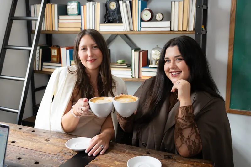 McKenzie and Sallie clinking latte cups at a wooden table